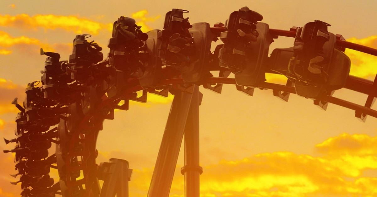 Silhouetted roller coaster train filled with riders traveling along a curved track against a dramatic golden-orange sunset sky.