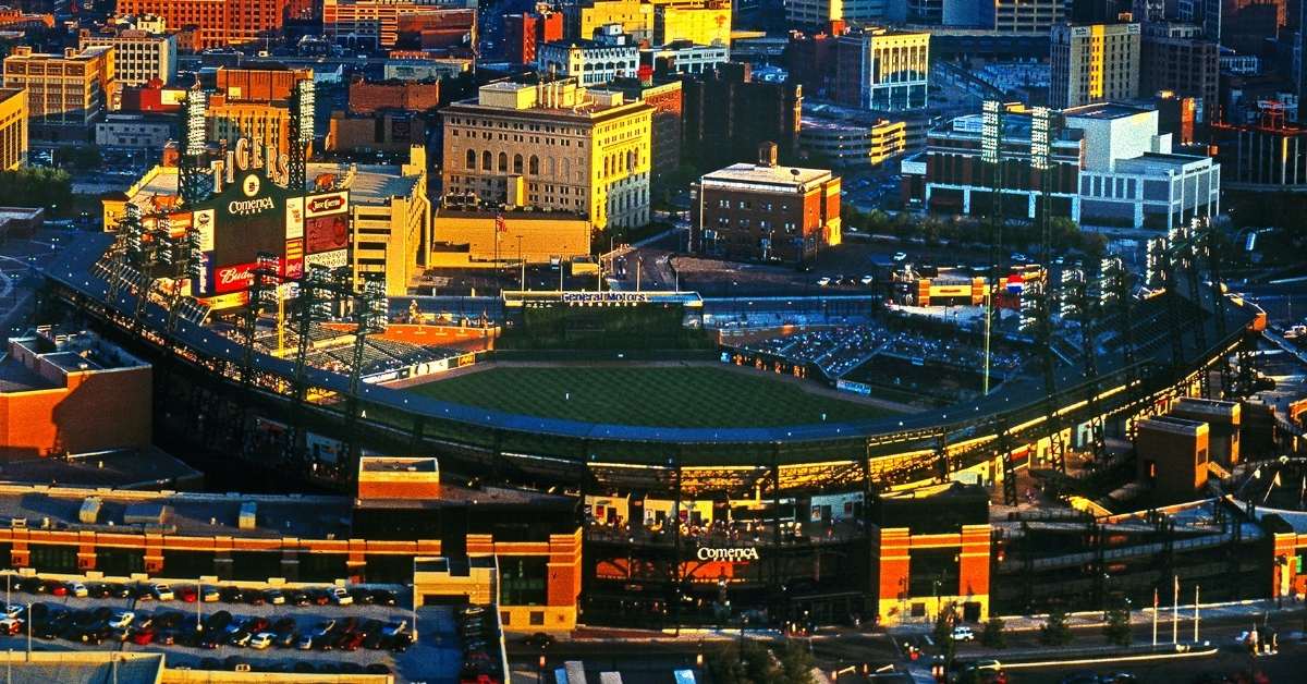 High-angle aerial photo of Comerica Park in downtown Detroit during golden hour, showing the Tigers baseball stadium, green field, packed seating, surrounding city buildings, and strong contrast between warm sunlight and cool shadows.
