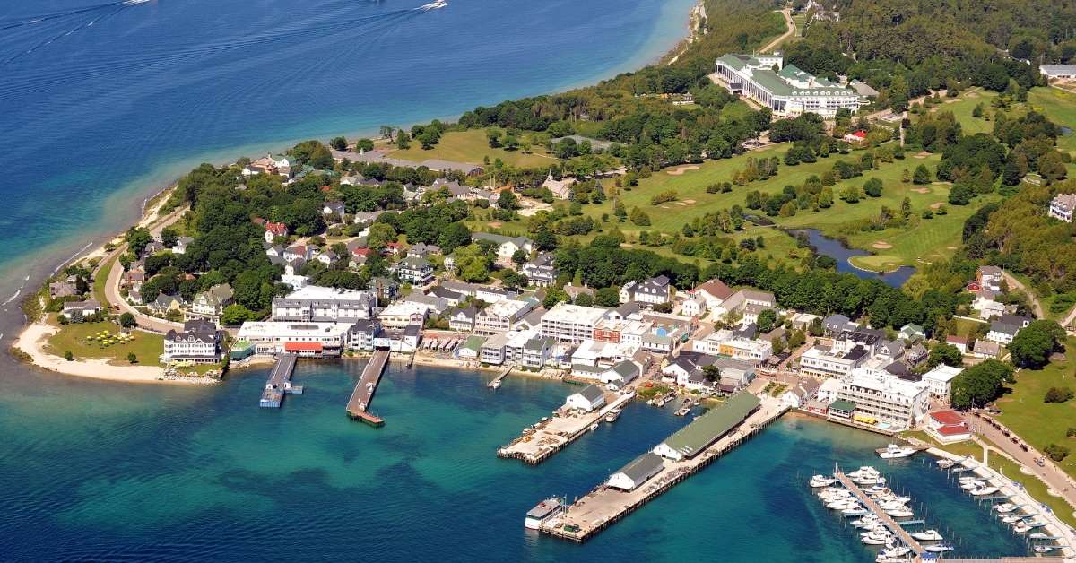 Aerial view of Mackinac Island, Michigan, showing turquoise harbor water, ferry docks, a marina full of boats, historic waterfront buildings, and the Grand Hotel on a green hillside.