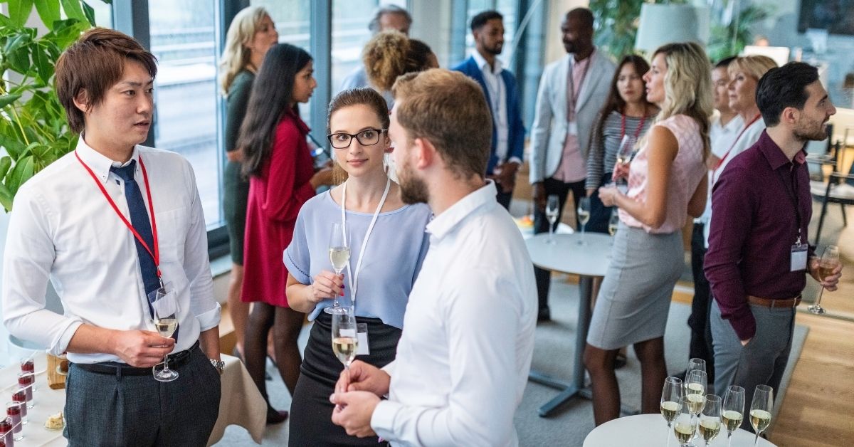 Professionals wearing conference badges chat and hold wine glasses during a networking event in a bright modern office venue.